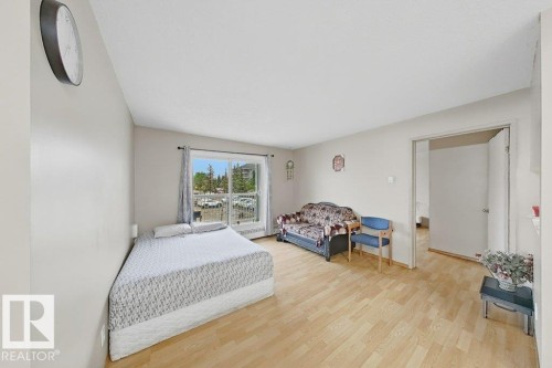 Spacious room featuring light wood-finish flooring, a bright window with a white balcony railing, and a doorway opening to another area - 218 5065 31 Avenue, Edmonton, AB 