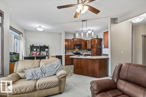 Open-concept living area featuring a ceiling fan with integrated lighting, soft grey carpeting, and a kitchen with wood-finish cabinetry, a peninsula island with counter seating, and a pendant light fixture - 229 41 Summerwood Boulevard, Sherwood Park, AB - Indoor Photo Showing Living Room