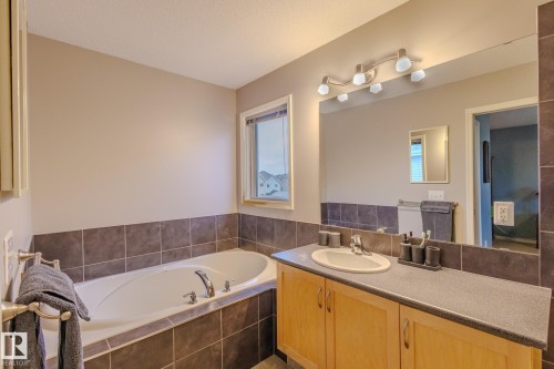 Bathroom featuring a built-in tub with tile surround, a single vanity with light wood-finish cabinetry, a large vanity mirror, and a window with a neutral frame - 7207 14A Avenue, Edmonton, AB - Indoor Photo Showing Bathroom