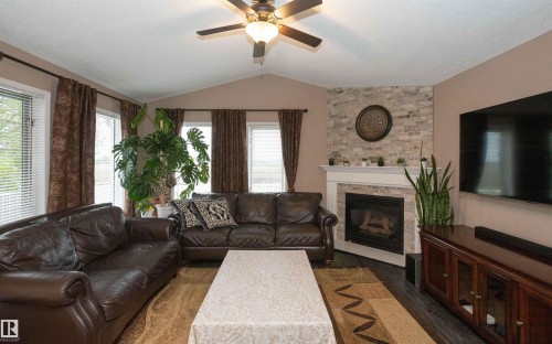 Living room featuring a vaulted ceiling with a ceiling fan, a stone-accented fireplace with a white mantel, multiple windows with blinds, and dark wood-finish flooring - 10739 109 Avenue, Westlock, AB 