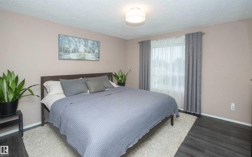 Spacious bedroom featuring dark wood-finish flooring, a window with sheer and blackout curtains, light neutral wall paint, and a ceiling-mounted light fixture - 10739 109 Avenue, Westlock, AB 