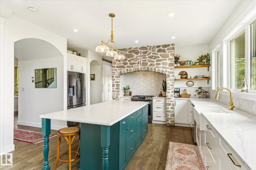 Spacious kitchen featuring a central island with a white countertop and teal cabinetry, a stone-clad range hood, white shaker-style perimeter cabinets, and a white farmhouse sink with a gold-finish faucet - 59102 Rge Road 234, Rural Westlock County, AB 