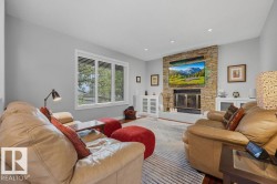 Living room featuring a stacked stone wood burning fireplace with an integrated media niche, hardwood flooring, and a large window with white louvered blinds - 