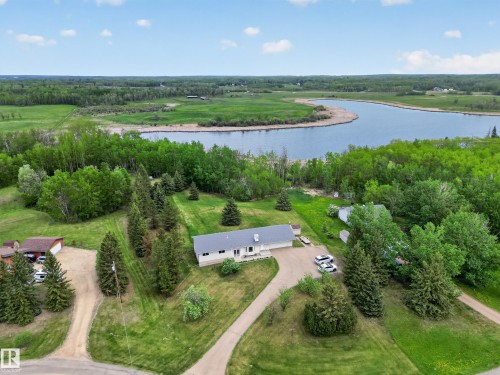 Expansive property featuring a single-story residence with a gray shingle roof and white siding, set against a backdrop of mature trees and a substantial body of water - 110 52308 Rr 214, Rural Strathcona County, AB 
