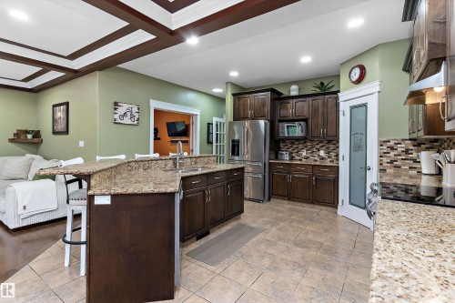 Open-concept kitchen featuring an eat-at island with a sink, granite countertops, wood cabinetry, stainless steel appliances, and a tiled backsplash - 16609 58A Street, Edmonton, AB 