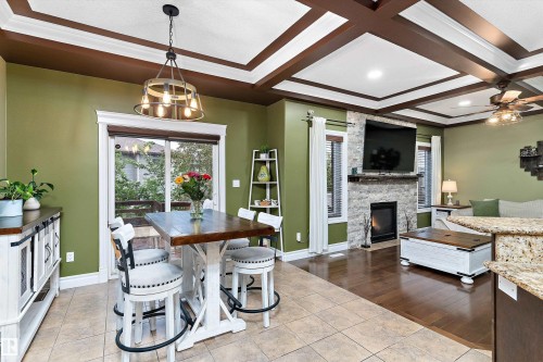Open-concept living space featuring a coffered ceiling, a stone-clad fireplace with a mantel, a sliding glass door, and a multi-light chandelier - 16609 58A Street, Edmonton, AB 