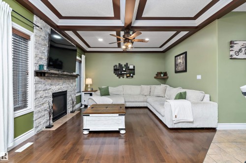 Living area featuring wood-finish flooring, a stacked stone fireplace with wood mantel, a coffered ceiling with a ceiling fan, windows with blinds, and light green wall paint - 16609 58A Street, Edmonton, AB 
