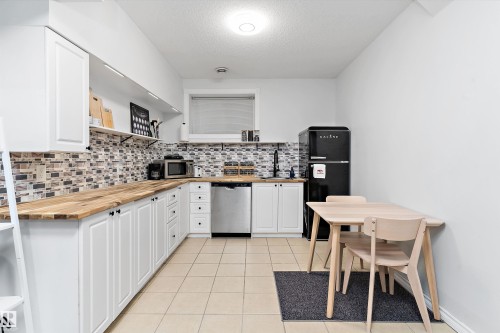 Contemporary kitchen featuring white cabinetry, wood-finish countertops, a mosaic tile backsplash, and stainless steel appliances including a dishwasher - 16609 58A Street, Edmonton, AB 