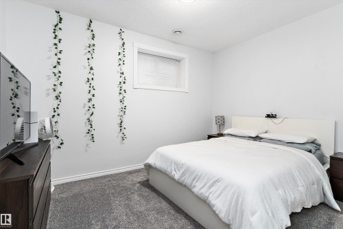 Bright room featuring a white headboard, a single window with blinds, dark wood-finish nightstands, and textured gray carpet flooring - 16609 58A Street, Edmonton, AB 