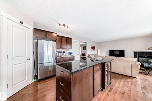 Open-concept living area featuring wood-finish flooring, a kitchen island with a dark countertop, dark wood cabinetry, a stainless steel refrigerator, and a fireplace with a dark tile surround - 2808 36 Avenue, Edmonton, AB 