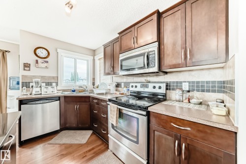 Kitchen featuring wood-finish flooring, dark wood cabinetry, stainless steel appliances, a corner sink with a window, and a tiled backsplash - 2808 36 Avenue, Edmonton, AB 