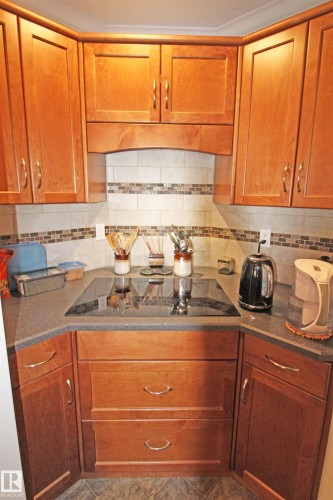Corner kitchen featuring wood cabinetry, an integrated cooktop, stone-finish countertops, a decorative tile backsplash, and tile flooring - 334 7001 Northview Drive, Wetaskiwin, AB - Indoor Photo Showing Kitchen