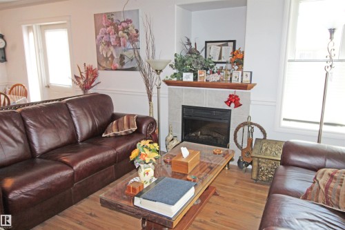 Living area featuring a fireplace with a tiled surround and wood mantle, wood-finish flooring, white walls with wainscoting, and large windows with blinds - 334 7001 Northview Drive, Wetaskiwin, AB - Indoor Photo Showing Living Room With Fireplace