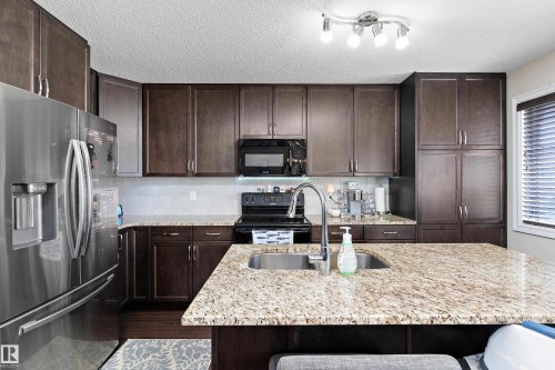 Kitchen featuring an island with an undermount double basin sink, granite countertops, stainless steel appliances, and dark wood-finish cabinetry - 6822 Cardinal Link, Edmonton, AB 