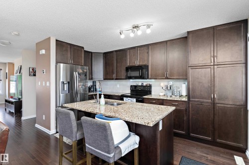 Kitchen featuring dark wood cabinetry, granite countertops, an island with a double basin sink, stainless steel refrigerator, and wood-finish flooring - 6822 Cardinal Link, Edmonton, AB 