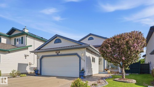 Light-colored siding exterior featuring a prominent attached garage with a white door, a paved driveway, and a manicured front lawn with a flowering tree - 856 Graham Wynd, Edmonton, AB 