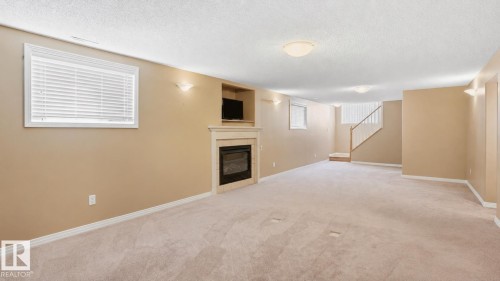 Expansive carpeted room featuring a built-in fireplace with a mantel, recessed entertainment nook, three flush-mount ceiling lights, and two wall sconces - 856 Graham Wynd, Edmonton, AB 