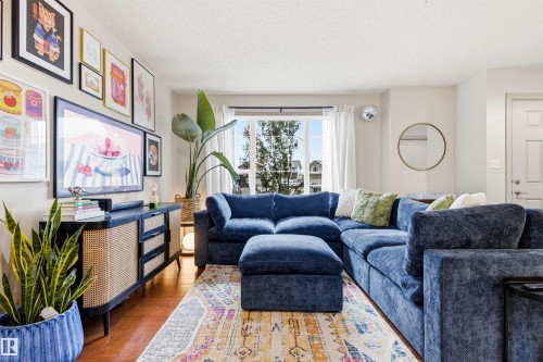 Living area featuring wood-finish flooring, a large window with white curtains, a built-in media console with woven cane and black paneling, a wall-mounted television, and a neutral color palette - 14807 141 Street, Edmonton, AB 