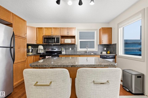 Full kitchen featuring wood-finish cabinetry, stainless steel appliances, a dark stone-finish backsplash, and a large central island with a stone-finish countertop - 14807 141 Street, Edmonton, AB 