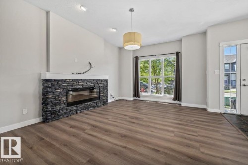 Living area featuring wood-finish flooring, an electric fireplace with a stacked stone facade and white mantel, a large picture window with dark drapes, and a ceiling-mounted light fixture - 356 Pioneer Road, Spruce Grove, AB 