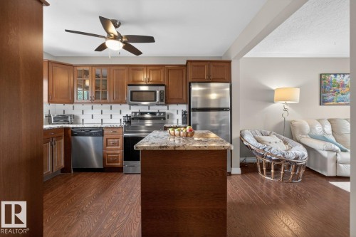 Open-concept kitchen featuring wood-finish flooring, a central island with a stone countertop, stainless steel appliances, wood cabinetry, and a tile backsplash - 9815 68 Avenue, Edmonton, AB - Indoor Photo Showing Kitchen With Stainless Steel Kitchen