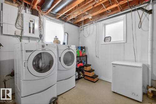 Dedicated laundry area featuring a white front-loading washer and dryer, utility sink, a chest freezer, and exposed wooden ceiling joists - 9815 68 Avenue, Edmonton, AB - Indoor Photo Showing Laundry Room