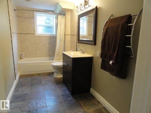 Bathroom featuring slate-look floor tiles, a white bathtub, a toilet, a dark wood-finish vanity with a light-colored countertop, and a wall-mounted mirror with an ornate light fixture - 9815 68 Avenue, Edmonton, AB - Indoor Photo Showing Bathroom