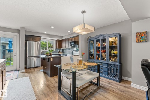 Open concept living area featuring wood-finish flooring, a contemporary kitchen with stainless steel appliances, a center island with seating, and a glass-top dining table beneath a modern pendant light - 5813 63 Street, Beaumont, AB 