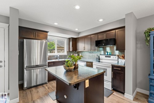 Kitchen featuring a central island with a stone-finish countertop, wood-finish flooring, dark wood cabinetry, stainless steel refrigerator, and white range - 5813 63 Street, Beaumont, AB 