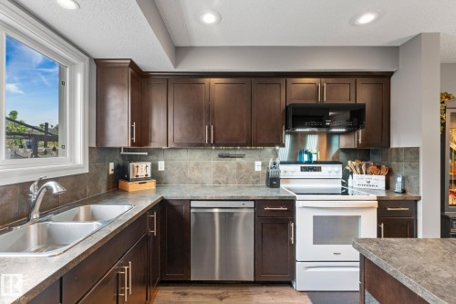 Kitchen featuring dark wood cabinetry, a stainless steel double basin sink, a stainless steel dishwasher, a white electric range with an overhead microwave, and stone-look countertops with a matching tile backsplash - 5813 63 Street, Beaumont, AB 