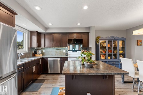 Spacious kitchen featuring an island with a stone-finish countertop, dark wood cabinetry, stainless steel appliances, wood-finish flooring, and recessed lighting - 5813 63 Street, Beaumont, AB 