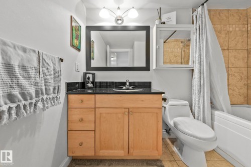 Bathroom featuring a light wood-finish vanity with a black countertop and integrated sink, a rectangular framed mirror, an overhead vanity light fixture, and a tiled shower/tub combination - 303 10165 113 Street, Edmonton, AB - Indoor Photo Showing Bathroom