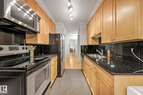 Galley kitchen featuring natural wood cabinetry, black granite-look countertops, and stainless steel appliances - 303 10165 113 Street, Edmonton, AB - Indoor Photo Showing Kitchen With Double Sink