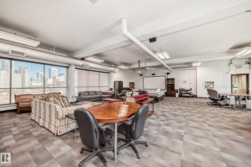 Expansive room featuring a wall of windows, geometric pattern carpet flooring, a ceiling-mounted projector screen, fluorescent lighting, and visible city buildings in the distance - 706 10125 109 Street, Edmonton, AB 