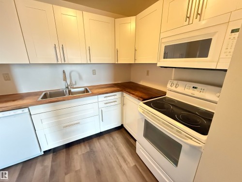 Kitchen featuring white shaker-style cabinetry, wood-finish countertops, stainless steel double-basin sink with a gooseneck faucet, white electric range, and wood-finish flooring - 706 10125 109 Street, Edmonton, AB 