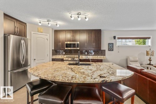 Open-concept kitchen featuring an expansive granite island, rich wood cabinetry, stainless steel appliances, and a dark tile backsplash - 1086 Grantham Drive, Edmonton, AB 