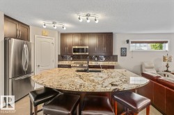 Open-concept kitchen featuring an expansive granite island, rich wood cabinetry, stainless steel appliances, and a dark tile backsplash - 