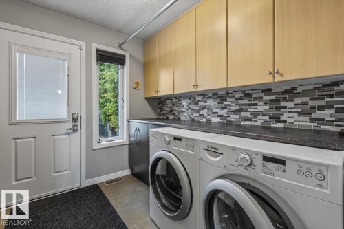 Functional laundry area featuring a full-light exterior door, a tall window, light wood-finish cabinetry, a patterned tile backsplash, and dark countertops - 1086 Grantham Drive, Edmonton, AB 