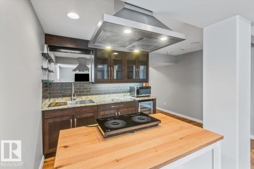 Modern kitchen with an induction cooktop island, granite countertops, dark wood-finish cabinetry, subway tile backsplash, and a stainless steel range hood - 1086 Grantham Drive, Edmonton, AB 