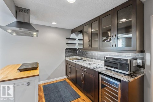 Modern kitchen featuring granite countertops, a subway tile backsplash, dark wood-finish cabinetry with glass inserts, wood-finish flooring, and a stainless steel range hood - 1086 Grantham Drive, Edmonton, AB 