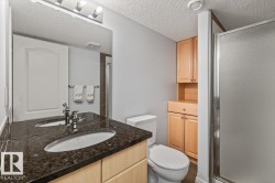 Bathroom featuring a dual sink vanity with a dark speckled countertop, light wood-finish cabinetry, and a walk-in shower with frosted glass enclosure - 