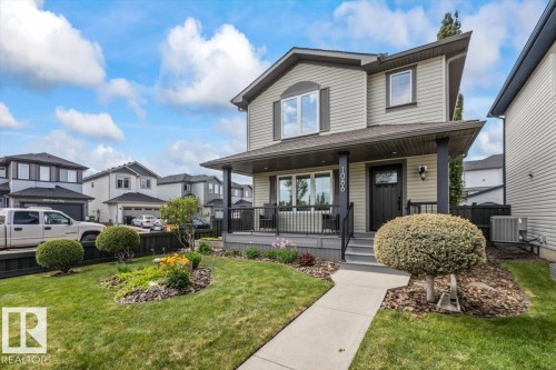 Modern two-story home featuring light-toned horizontal siding, a covered front porch with dark railings and columns, a landscaped front yard with a concrete walkway, and an arched architectural accent above the main window - 1086 Grantham Drive, Edmonton, AB 