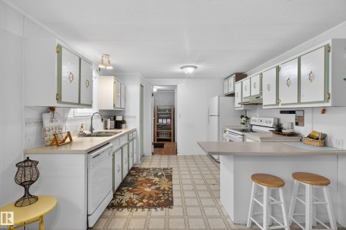 Kitchen featuring extensive white cabinetry with sage green accents and gold-tone hardware, light-colored countertops, a double basin sink with a gooseneck faucet, a white dishwasher, and a breakfast bar - 21 Willowdale Drive, Edmonton, AB - Indoor Photo Showing Kitchen With Double Sink