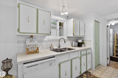 Kitchen featuring white cabinetry with green trim, a double basin sink with a gooseneck faucet, a white dishwasher, light-toned countertops, and a tiled backsplash - 21 Willowdale Drive, Edmonton, AB - Indoor Photo Showing Kitchen With Double Sink