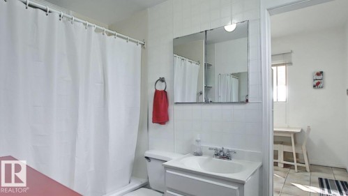 Bathroom featuring a single vanity with a white countertop, a medicine cabinet with integrated lighting, white tiled walls, a white toilet, and a shower with a white curtain - 10017 113 Street, Edmonton, AB 