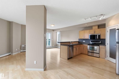 Open-concept living space featuring wood-finish flooring, a kitchen with light wood cabinetry, dark countertops, stainless steel appliances, and a dark tile backsplash - 901 10504 99 Avenue, Edmonton, AB 