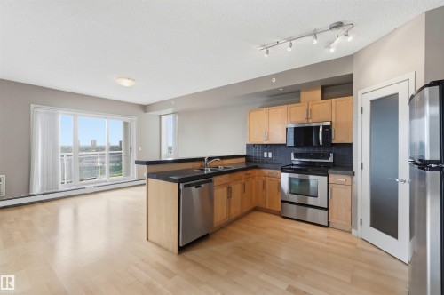 Open-concept kitchen featuring light wood cabinetry, stainless steel appliances, dark countertops, and a dark tile backsplash - 901 10504 99 Avenue, Edmonton, AB 