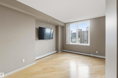 Spacious room featuring wood-finish flooring, a large window with city buildings visible, a wall-mounted television, neutral wall paint, and ceiling-mounted lighting - 901 10504 99 Avenue, Edmonton, AB 