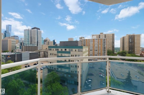 Balcony with clear glass railings overlooking a cityscape featuring various high-rise buildings and green tree canopies - 901 10504 99 Avenue, Edmonton, AB 