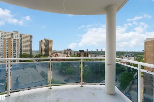 Outdoor balcony featuring a white railing and frosted glass panels, offering an expansive urban landscape with multiple high-rise buildings and a distant tree line under a bright sky - 901 10504 99 Avenue, Edmonton, AB 